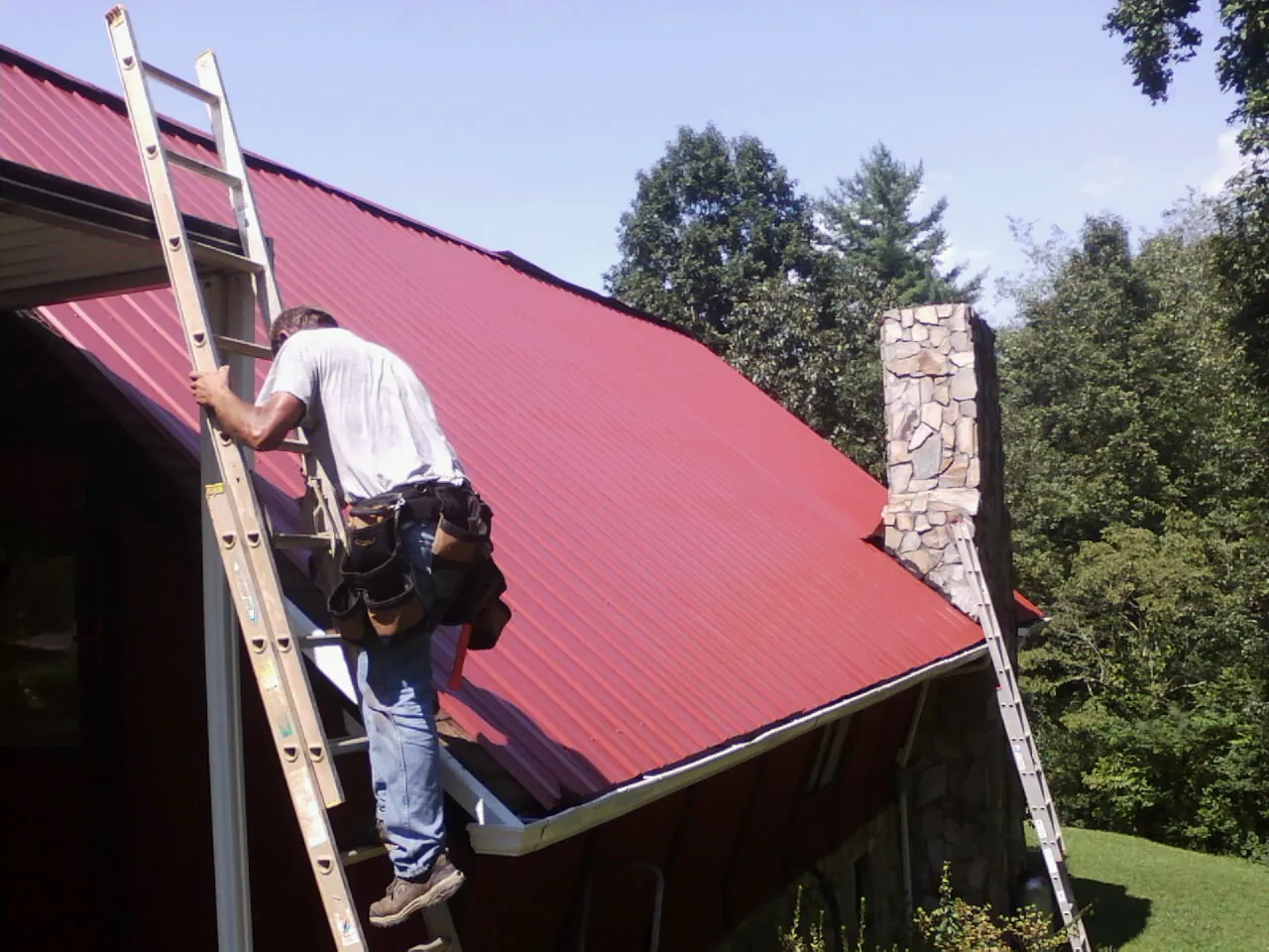 Roofer on a ladder beside a red metal roof and stone chimney — Soffit & Fascia Repair by Roofing Brothers in Trumbull
